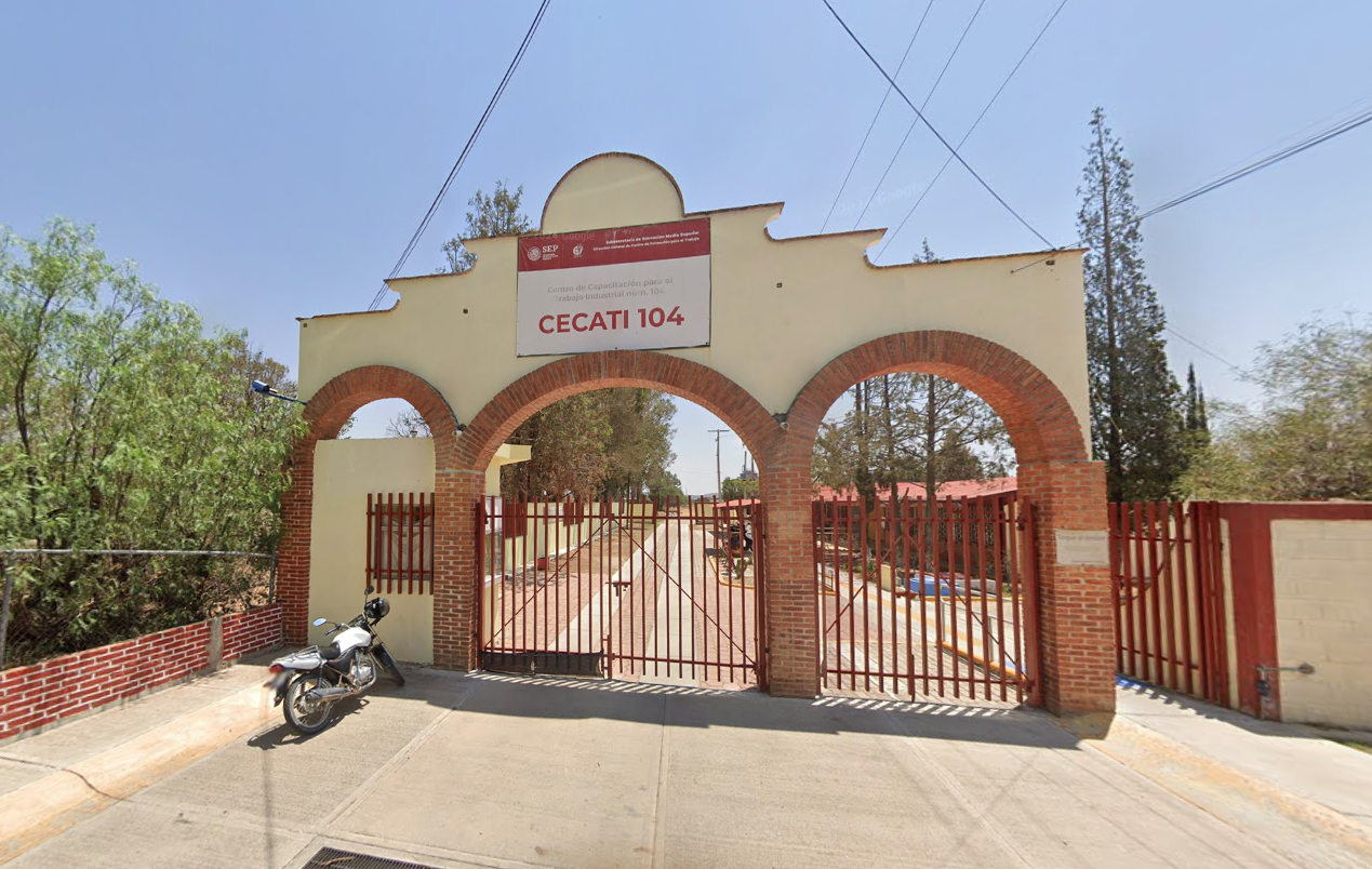 Fotografía en la que se muestra la entrada del plantel, conformada por tres arcos de tabique rojo. El arco de la izquierda tiene una caseta y los dos arcos de la derecha tienen rejas que dan acceso a las instalaciones. Arriba del arco central hay una lona que dice CECATI 104.